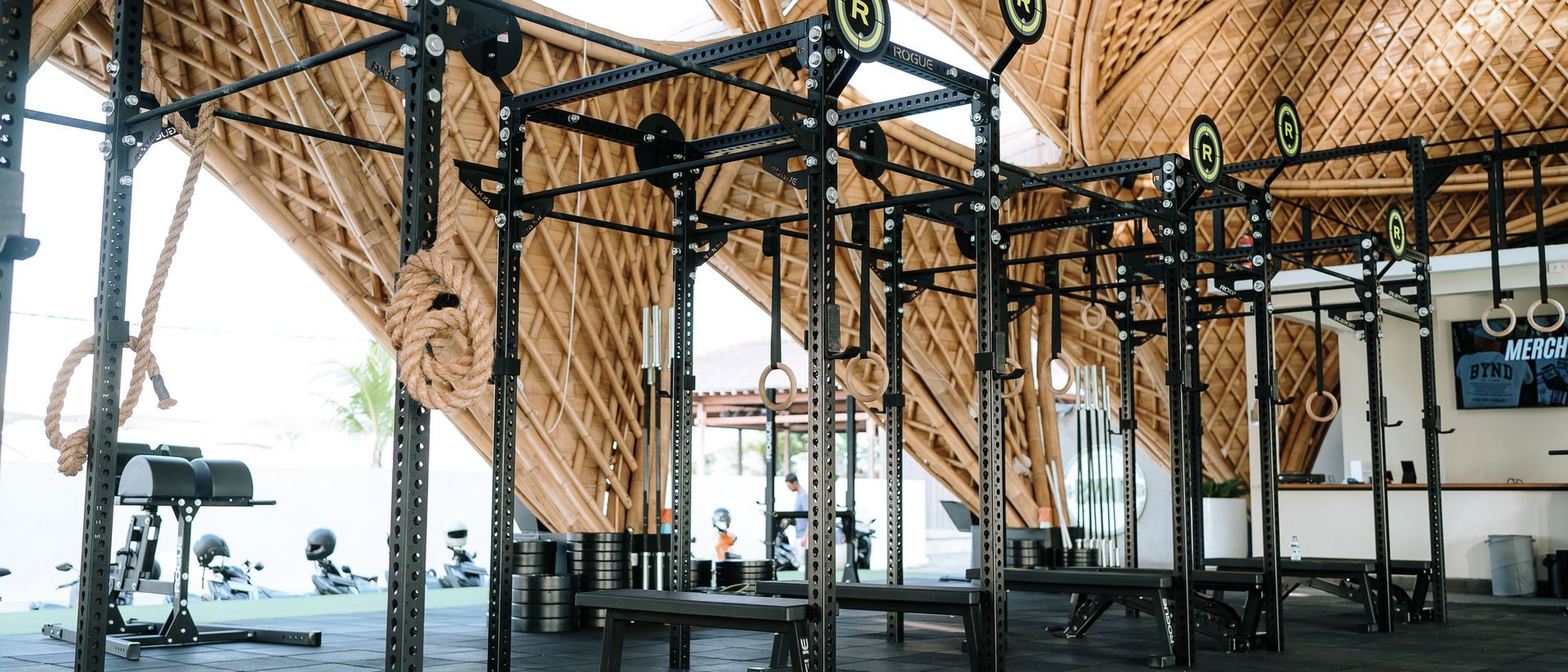 Steel squat rigs, benches and stacked weight plates under a curved bamboo roof at a partner gym in Bali