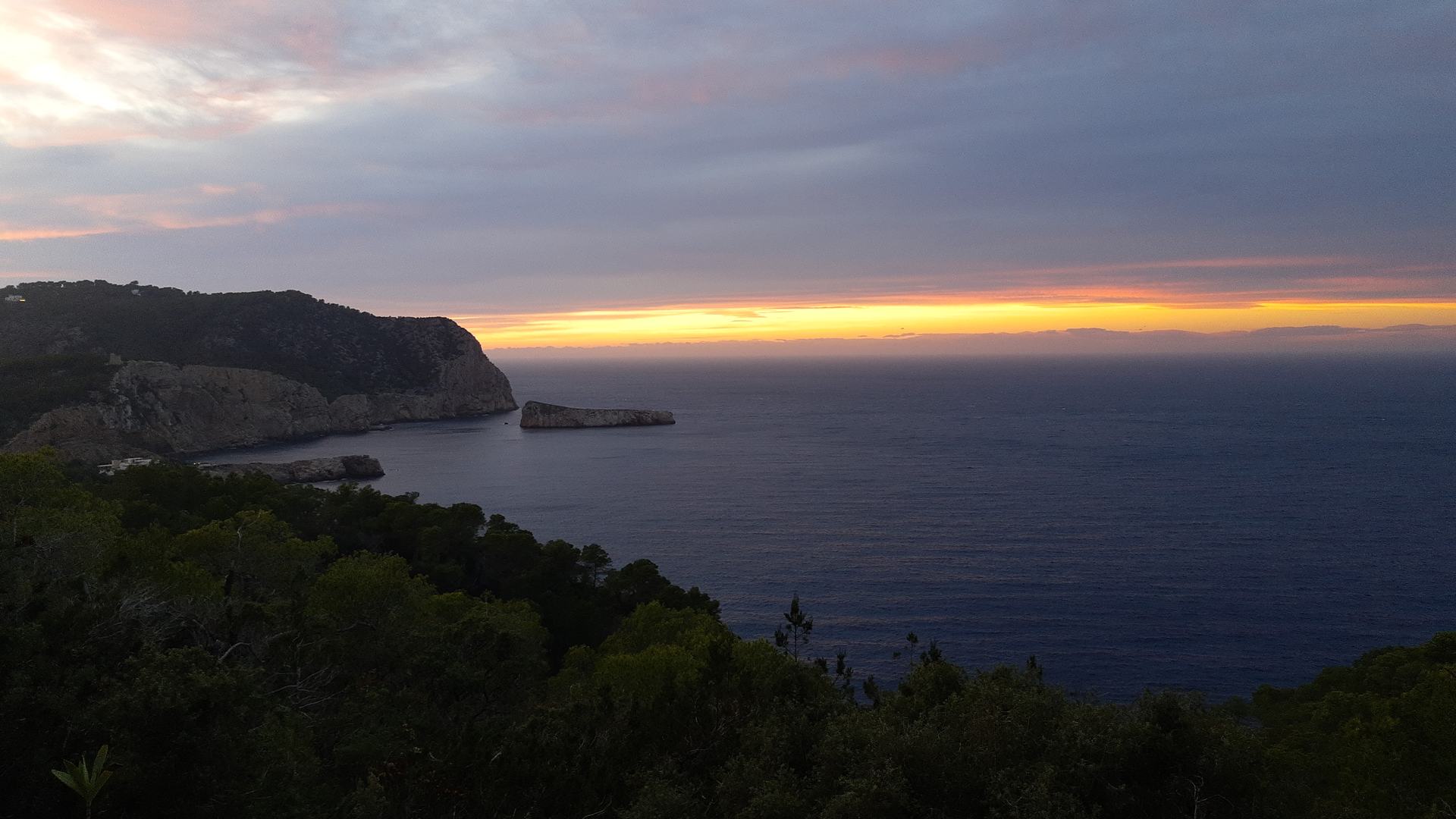 Sunset over the sea from the west coast of Ibiza, a wooded headland and a small islet in silhouette