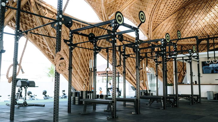 Steel squat rigs, lifting benches and stacked weight plates on a black rubber floor, under a curved bamboo roof open to the fields.