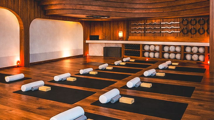 A dark timber studio with mats, bolsters and blocks laid out in rows, and dumbbells, bars and plates racked along the back wall.