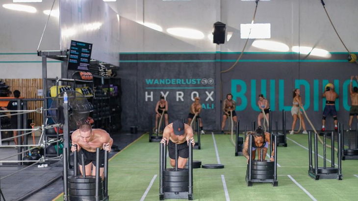 Weighted sleds lined up on green turf lanes in a warehouse gym, with racked dumbbells and wall balls along the back wall.
