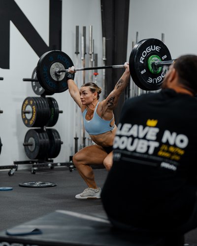 Kyra Milligan holding a barbell overhead at the bottom of a squat in a gym